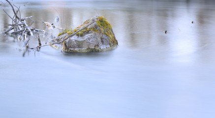 A single rock is on the edge of calm water in the morning at sunrise with plenty of space for text and water as a background