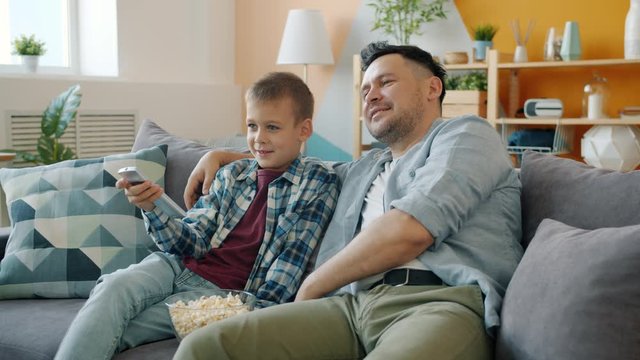 Little boy is watching TV at home with caring dad middle-aged man sitting on sofa in living room talking having fun together. Family leisure activities concept.