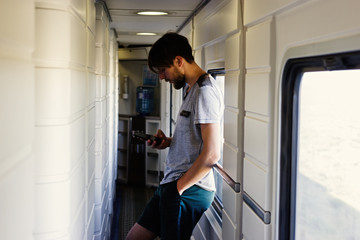 Handsome young caucasian man with a beard, looking at a smartphone in the hallway by the window . Travel leisure