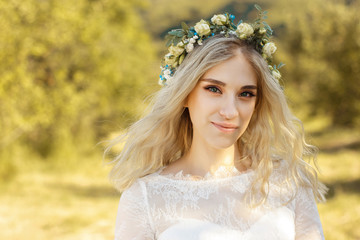 Beautiful blonde young bride portrait with flower bouquet and wreath on her head in white wedding dress outdoor in summer