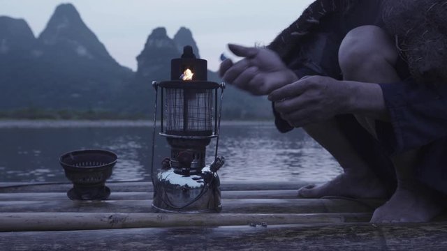 Close shot of a Chinese cormorant fisherman priming an oil lantern on a bamboo raft with water behind and karst towers in the bckground