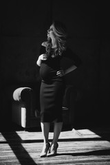 Beautiful young sexy pregnant woman with curls and make-up in a black tight-fitting dress. Black and white photo. Fine art. Waiting for a child. Soft selective focus.