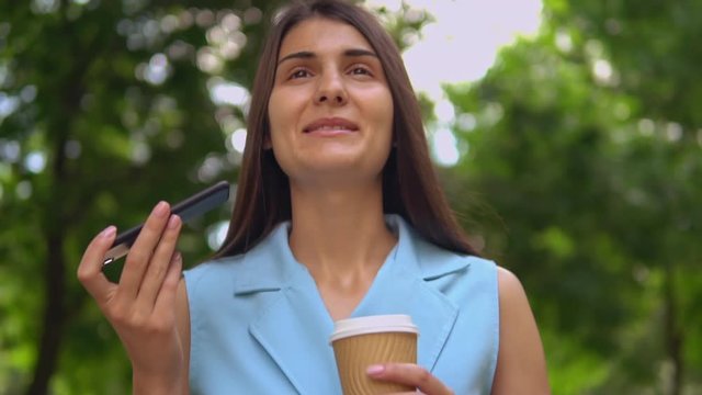 brunette walks in summer park student holding coffee to go using app on smartphone summer season in city slow motion
