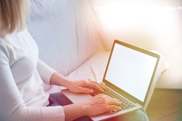 Naklejka premium Portrait of blonde hair woman using her blank screen laptop while sitting on the sofa at home in the sunset light. Blonde female sit indoors at home working on computer with blank screen. Home office 
