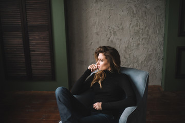 A beautiful woman with curls and full lips in a black turtleneck and jeans sits in an armchair in a loft style studio. Soft selective focus.