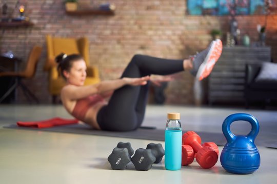 Focus on fitness equipments, barbell and kettlebell. Woman doing sit-ups in the background. Concepts about home workout, fitness, sport and health.