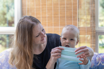 Mom is ready to puts on a disposable protective face mask to her little son. The baby with open mouth is looking at the camera.