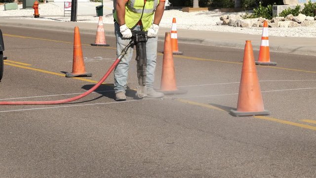 Working man uses a jackhammer on the asphalt of a city street next to the orange safety warning cones, as part of a utility project.