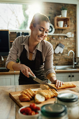 Young happy woman slicing baguette while preparing food in the kitchen.
