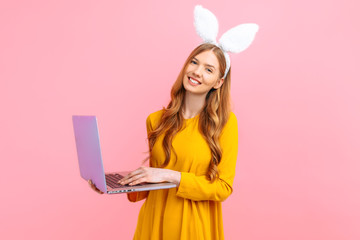 happy Easter. Happy woman in the ears of an Easter Bunny holding a basket of Easter eggs using a laptop on an pink background