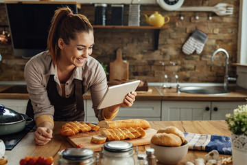 Happy woman reading recipe on touchpad while preparing food in the kitchen.