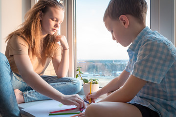 Children together sit on windowsill and draw with colored pencils..Entertainment and play for kids on quarantine indoor. Family hobbies at home. Safety during global infections and epidemics.