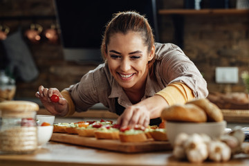 Happy woman adding cherry tomato while making bruschetta in the kitchen.