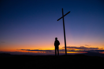a man near the cross stands on a mountain and looks at the sunset