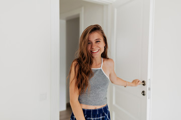 Laughing adorable girl with long wavy hair wearing gray top walks into the room and smiles at the camera