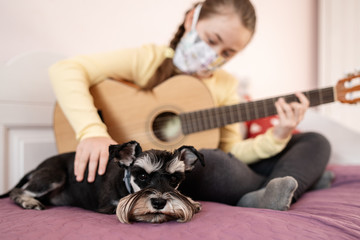 Young white girl in antibacterial mask playing her guitar sitting on the bed at home due to the isolation of COVID-19. Quarantine at home during the virus epidemic disease prevention. Puppy on bed.