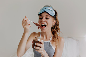 Happy charming young woman with sleeping mask wearing pajamas eating chocolate cream with teaspoon...