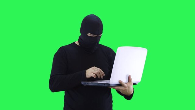 A man in a black mask enters data into a laptop, on a green background