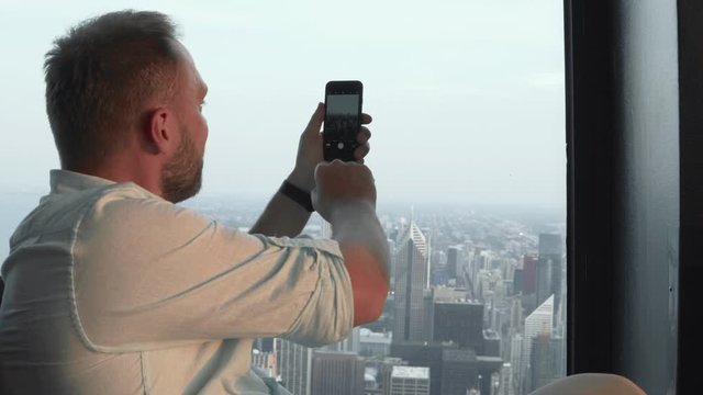 Young handsome man taking photos on smartphone. Male shoots panorama of Chicago, America from skyscraper.