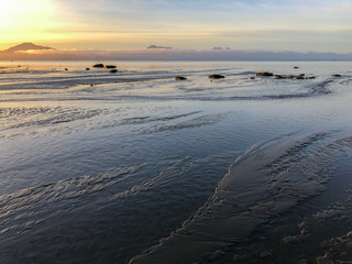 Water sheets of sandy beach as sunrise catchs three distance peaks rising about a fog bank