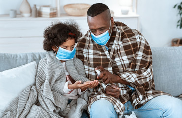 Epidemic hygiene strategies. African American family wearing protective masks and cleaning hands with sanitizer indoors