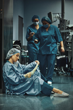 Tired Doctor Sitting After Surgery In The Hospital Corridor. Great Burden On Doctors And Medicine. Rest After Work. Epidemic, Lack Of Medical Personnel, Medical Wards.