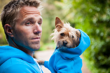 Dog owner carrying his scruffy little best friend in matching blue hoody sweatshirts standing outdoors in front of greenery