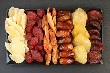 Сandied slices of ginger root and strawberries, dried pear slices, dates, peaches and apricots lying on a black ceramic plate on a black stone surface. Macro