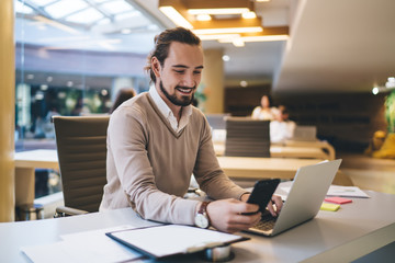Obraz premium Happy Caucasian man in smart casual wear checking received mobile message during remote work on modern netbook, successful freelancer using 4g wireless internet for connecting on smartphone device