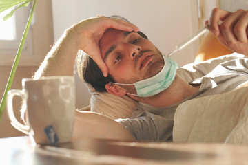 Coronavirus quarantine: young man lying in his bed with medical mask and thermometer