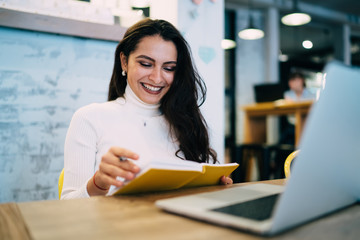 Fototapeta premium Cheerful woman with brunette hair reading notes from knowledge textbook preparing to college examination, happy female student studying paper information near table with laptop device for browsing