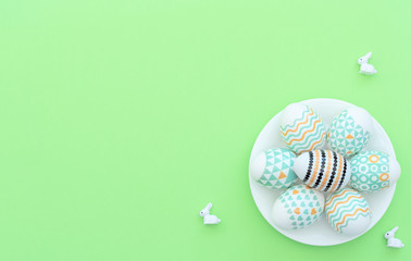 Decorated Easter egg lie on the white plate on green background with small bunnies. Happy Easter holiday concept. Greeting, invitation card. Flat lay style with copy space.