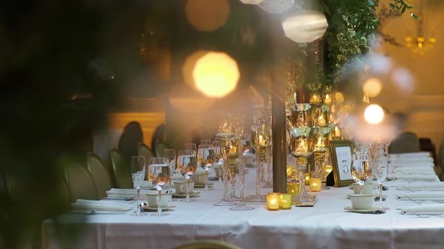 Square table served with wine glasses, cutlery and glassware, decorated with stylish candlesticks, and green branches with leaves