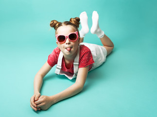 A teenager girl in pink sunglasses and a white sundress lies and shows her tongue.