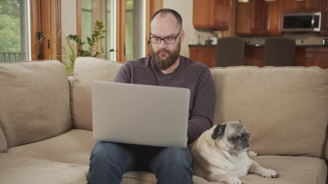 Candid lifestyle, hipster man with beard and glasses works from home sitting at laptop computer with cute pet pug dog while typing. Relaxed successful man online or working from home.