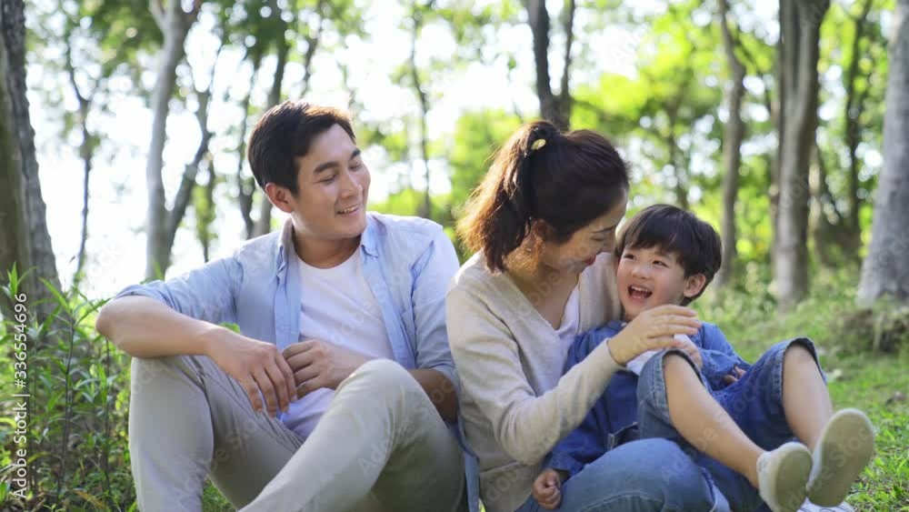 Wall mural young asian couple sitting on grass having fun with son outdoors in woods