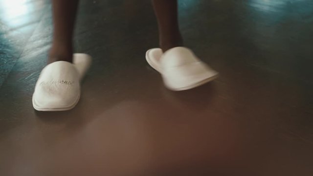 African woman is dancing dressed in white robe and white fluffy slippers, front view of dancing legs on the wooden floor in the room, morning preparation for the wedding