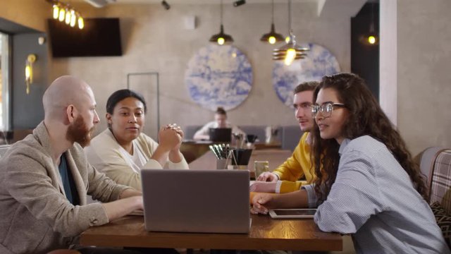 Diverse team of businessman and businesswomen sitting together at table in coffeeshop and discussing project on laptop screen