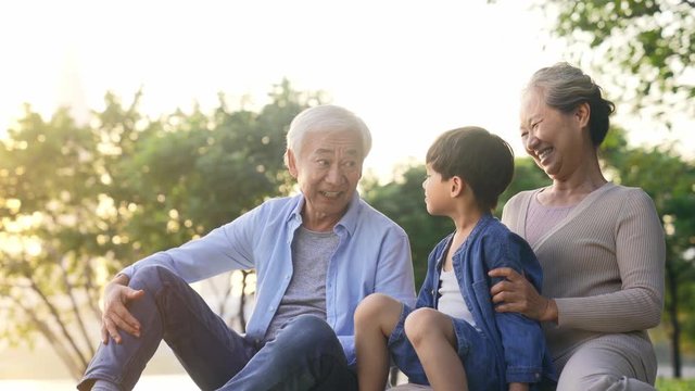 happy senior asian couple sitting on grass having fun with grandson outdoors in park