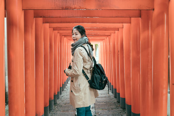 Fototapeta na wymiar side view of beautiful asian chinese woman walking through famous lines of orange column Torii at tenmangu temple in osaka. girl backpacker holding camera turn face around standing under red gates