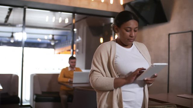 African american woman in casual clothes walking through cafe and surfing the Internet on digital tablet