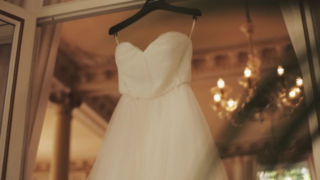 Beautiful tender white wedding dress is hanging on the window frame in the room, wedding preparation, bridal details