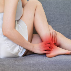 woman wearing white Sleepwear sitting on gray sofa . Female holding hand to spot of ankle-ache. Concept photo with read spot indicating location of the legs pain.Health care concept,Soft focus