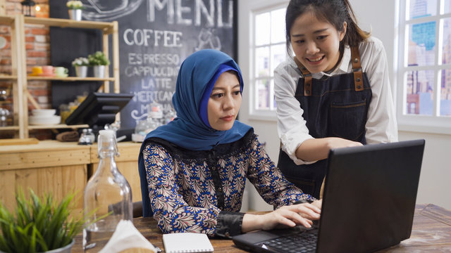 saudi business woman freelance working in cafe bar. friendly waitress friend pointing on laptop screen and having discussion with muslim worker in coffee house. islam female correction on keyboard