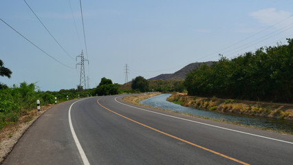 Fototapeta premium Irrigation and transportation system, Curve asphalt road along the canal with mountain and high voltage tower in Thailand
