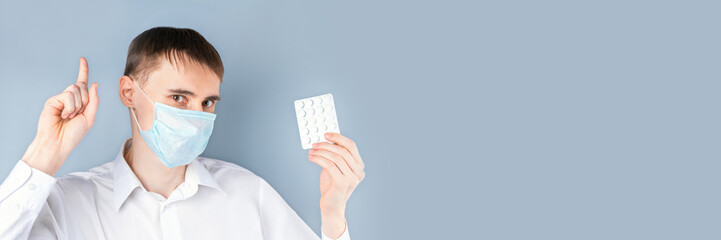Banner. A man in a medical mask holds pills  in one hand, the other shows the index finger up on a gray background, there is an empty space on the right for the text. Coronavirus, Covid-19