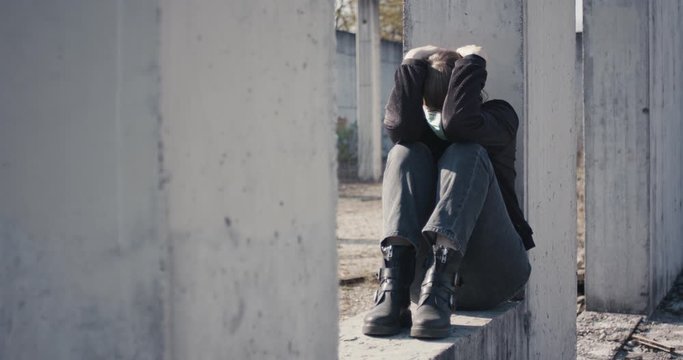 Woman with face mask full of anxiety and depression sitting at an empty abandoned space closed for safety isolation and social distance due to coronavirus covid 19 pandemic outbreak full shot