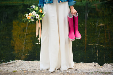 bride in white dress holding in hands a bouquet of flowers and pair of purple rubber boots 