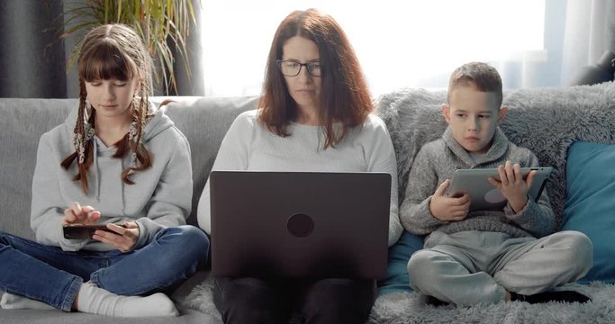 Mature woman in eyeglasses and domestic outfit working on laptop while her children using personal gadgets on couch. Modern family values.