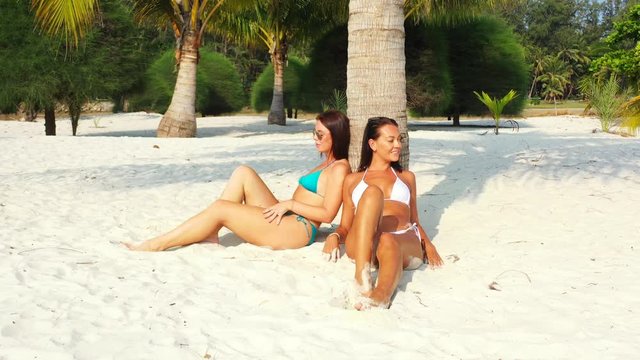 Two young women enjoying exotic beach with white sand sunbathing backed on a palm tree trunk in Bermuda
